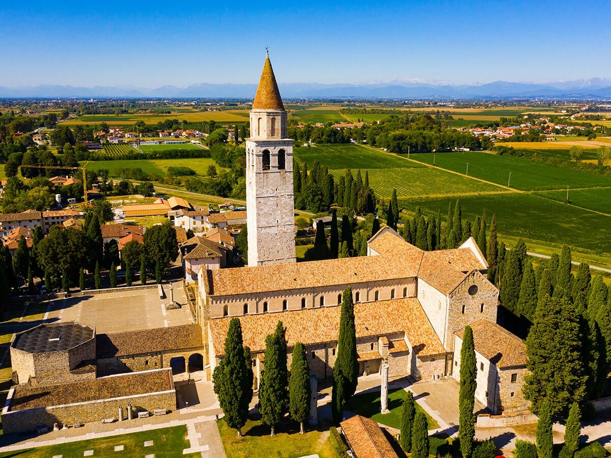 Luftaufnahme der Patriarchalbasilika von Aquileia mit ihrem Campanile, umgeben von grünen Feldern und Zypressen.