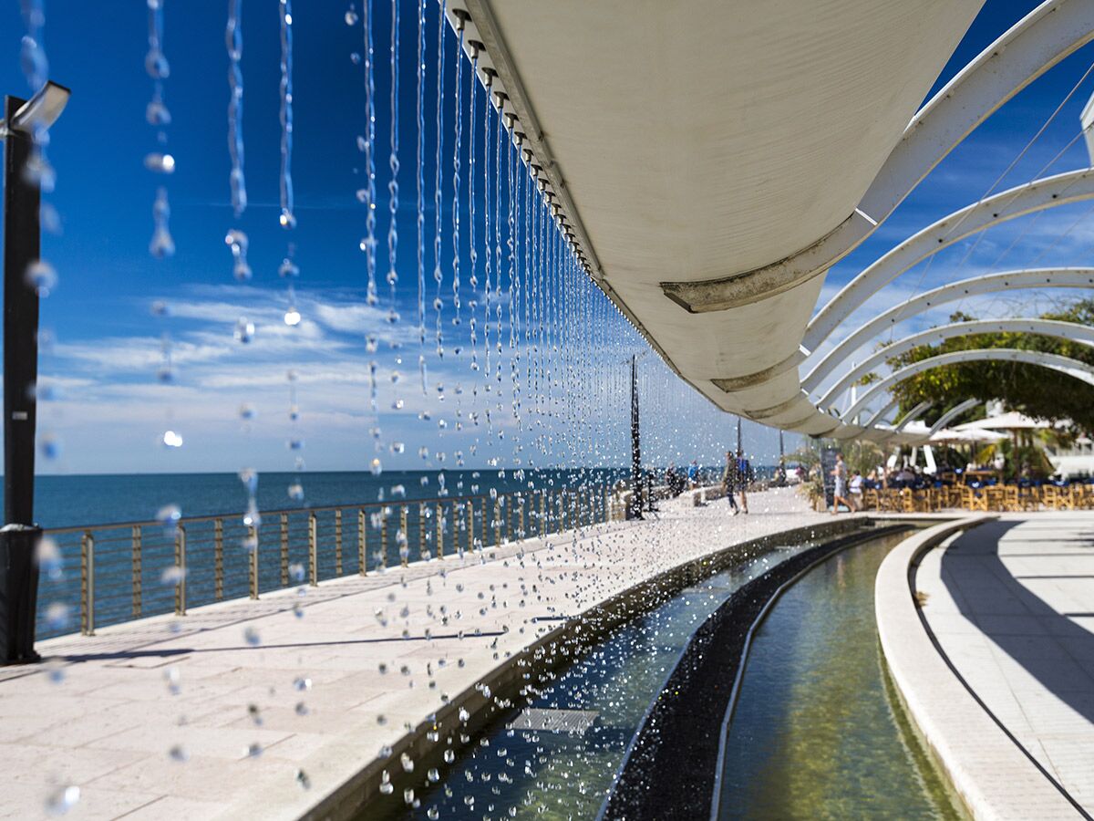 Moderner Wasservorhang an einer sonnigen Strandpromenade mit Blick auf das Meer, Menschen flanieren.