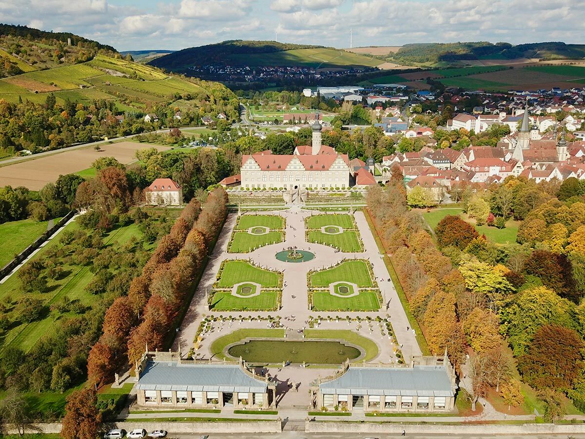 Luftaufnahme von Schloss Weikersheim mit seinem großen Barockgarten und Herbstbäumen, umgeben von Weinbergen und einer Stadt.
