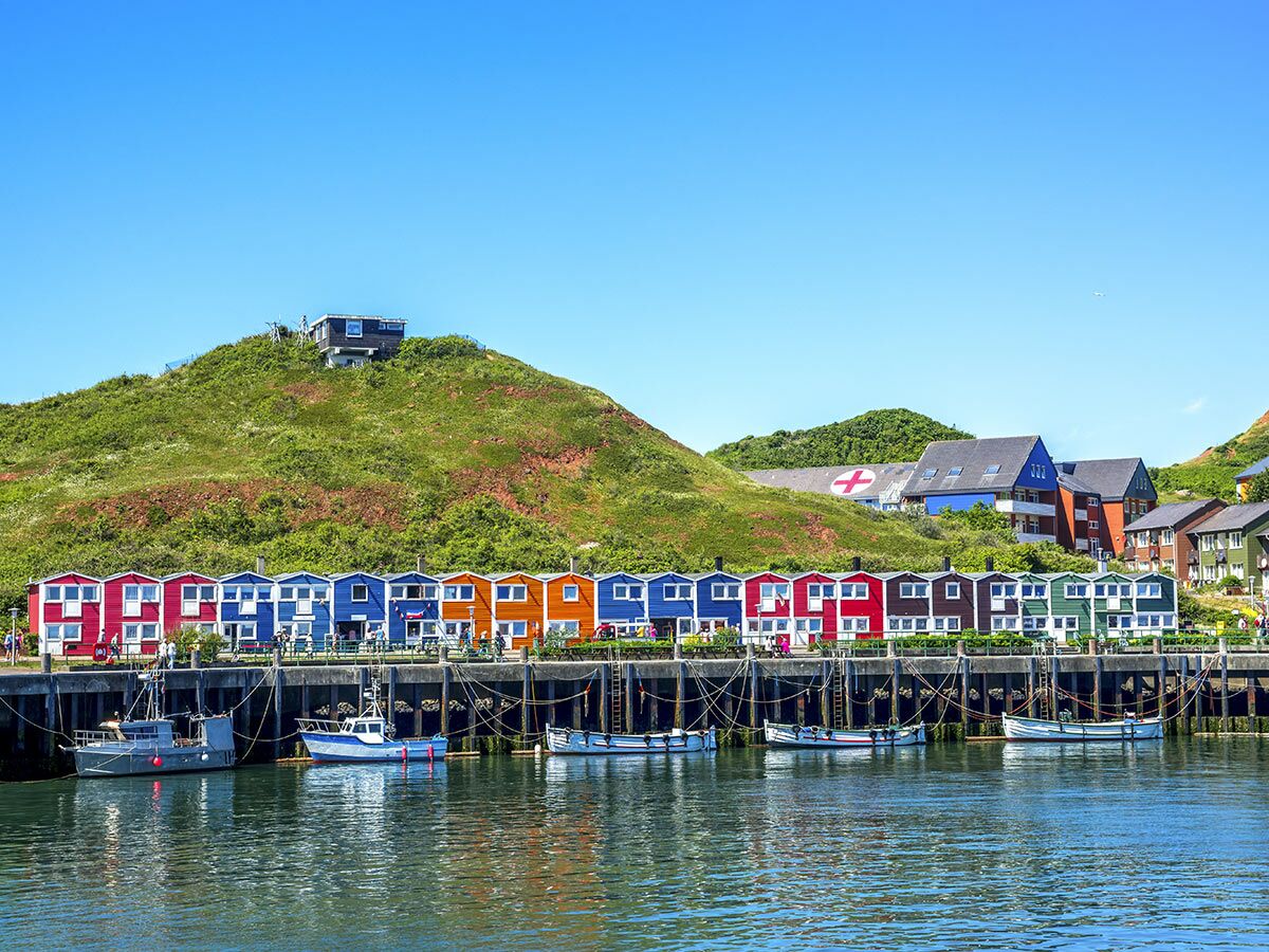 Farbenfrohe Fischerhäuser in einer Reihe am Hafen von Helgoland mit Fischerbooten davor, umgeben von grünen Hügeln unter blauem Himmel.