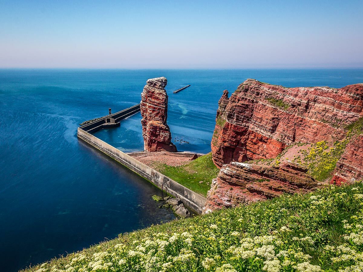 Dramatische rote Felsen und die Lange Anna auf Helgoland mit blauem Meer und einem Pier unter klarem Himmel.