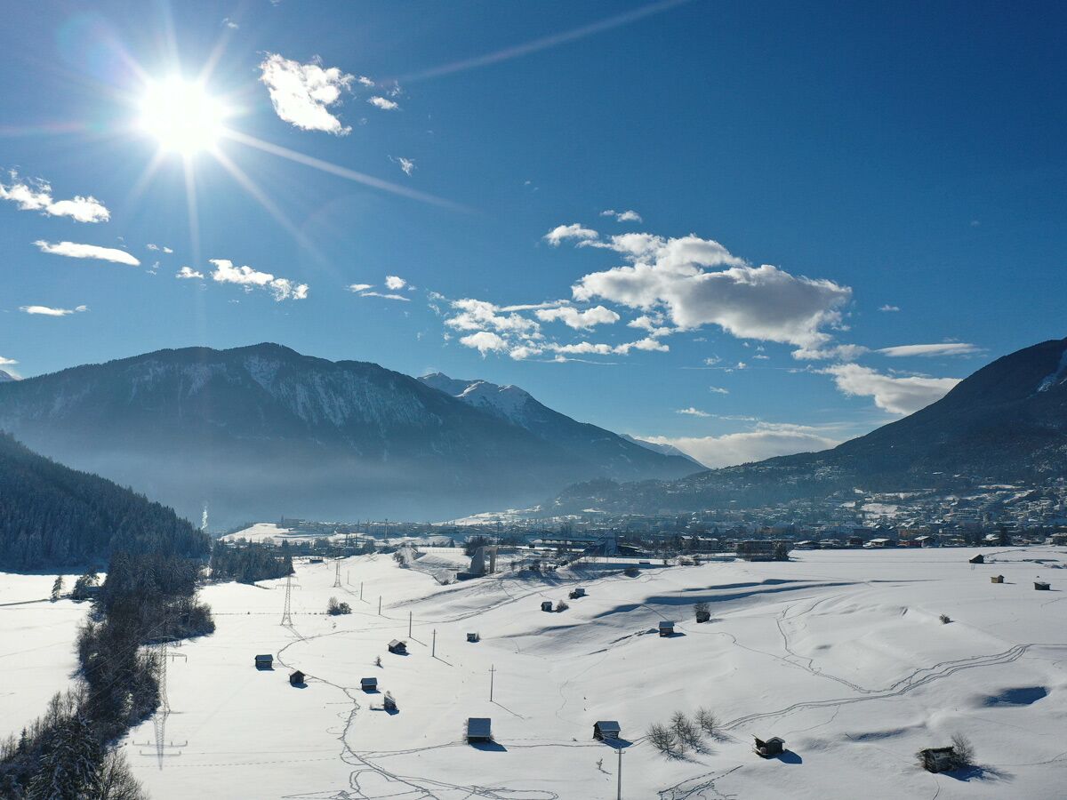 Sonnige Winterlandschaft in den Alpen mit schneebedeckten Bergen, Tal, Dorf, Hütten und Spuren im Schnee unter blauem Himmel.
