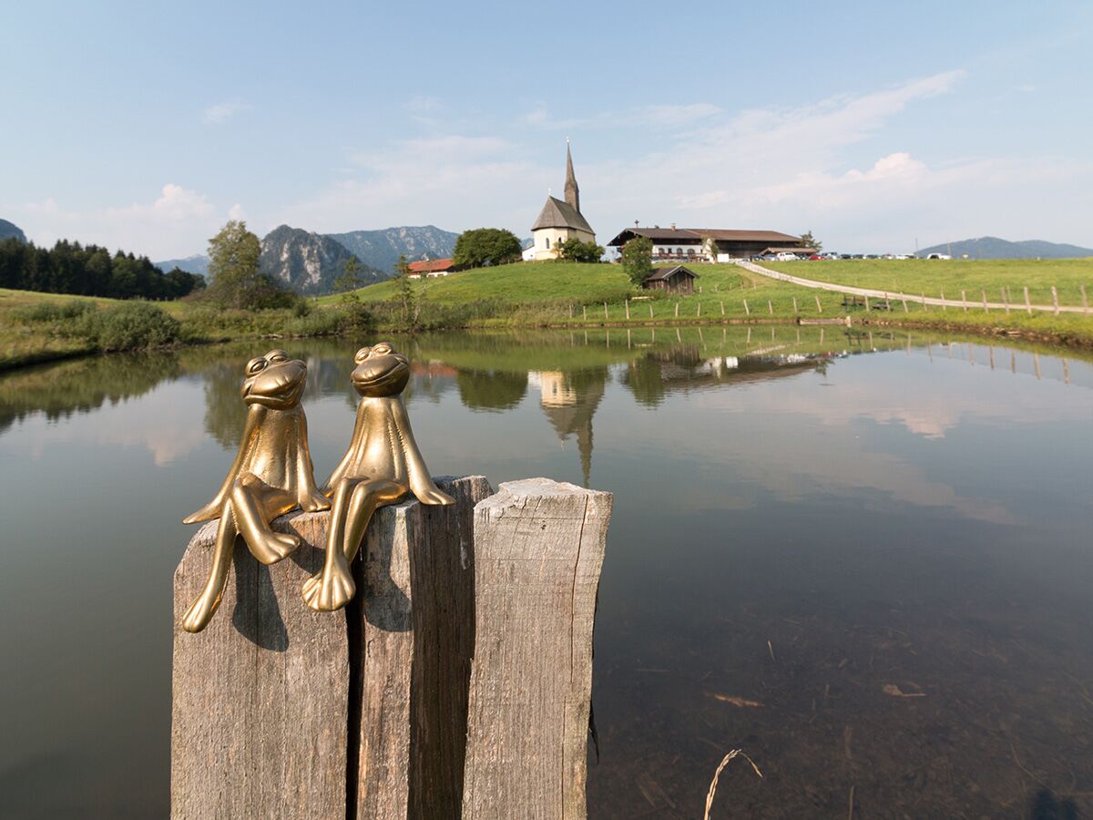 Zwei goldene Froschfiguren sitzen auf Holzpfählen an einem ruhigen See mit einer Kirche und Bergen im Hintergrund.