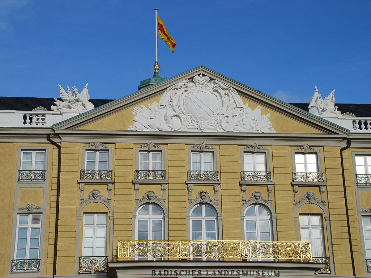 Gelbes Gebäude Badisches Landesmuseum Karlsruhe mit Wappen, Skulpturen, vielen Fenstern und gehisster gelb-roter Flagge unter blauem Himmel.
