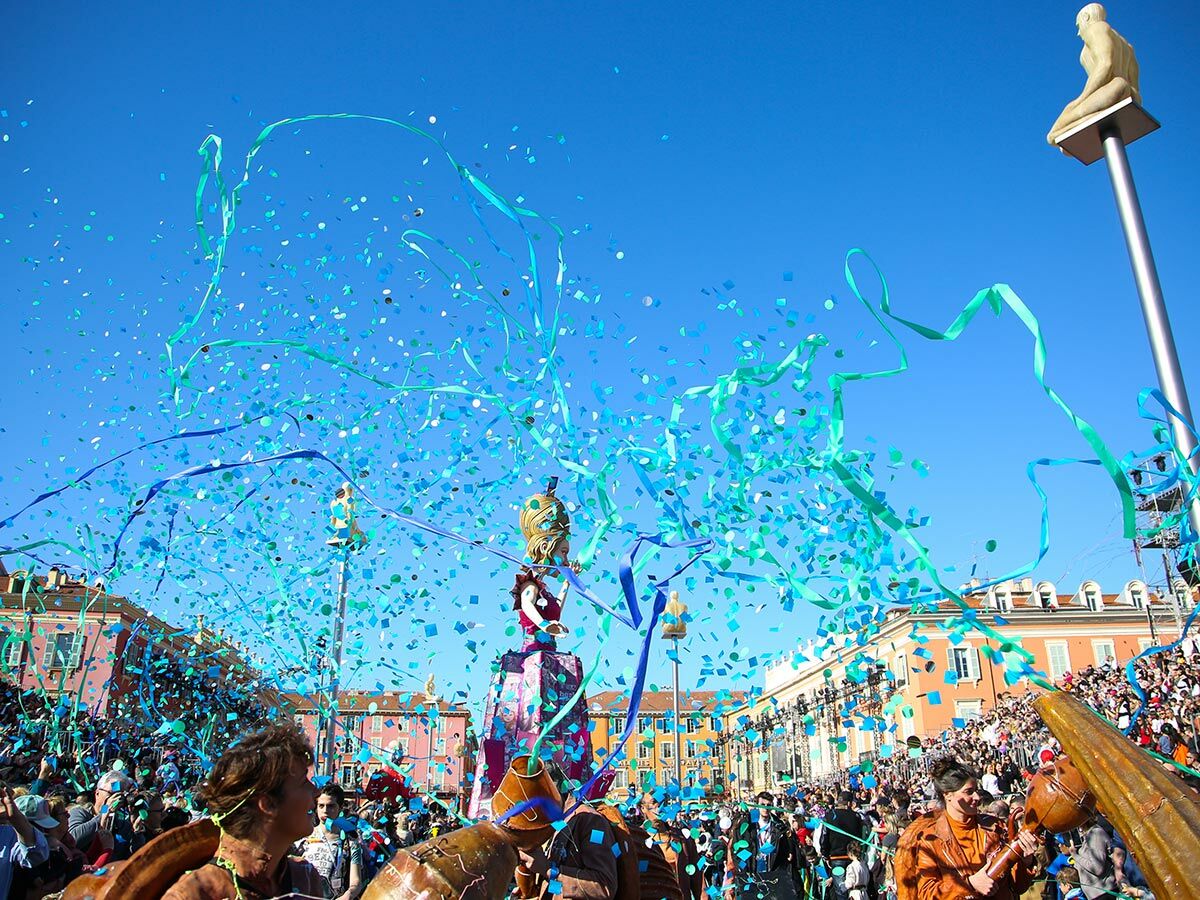 Nizza Karnevalsumzug: Blaues Konfetti und Bänder fliegen über jubelnde Menschen und bunte Gebäude unter blauem Himmel.