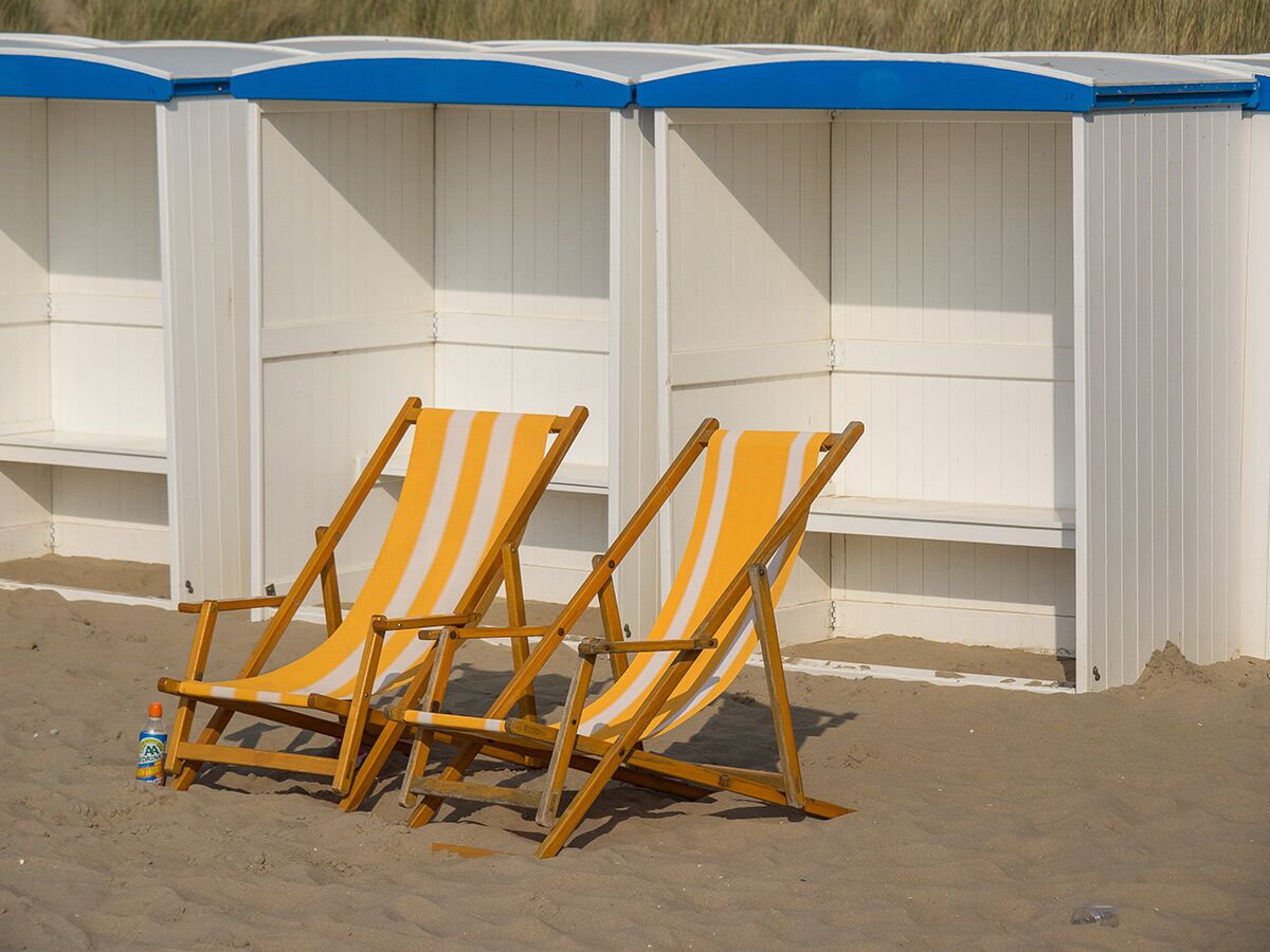Zwei gestreifte Strandstühle und weiße Strandhütten mit blauen Dächern auf einem Sandstrand, kleine Flasche daneben.