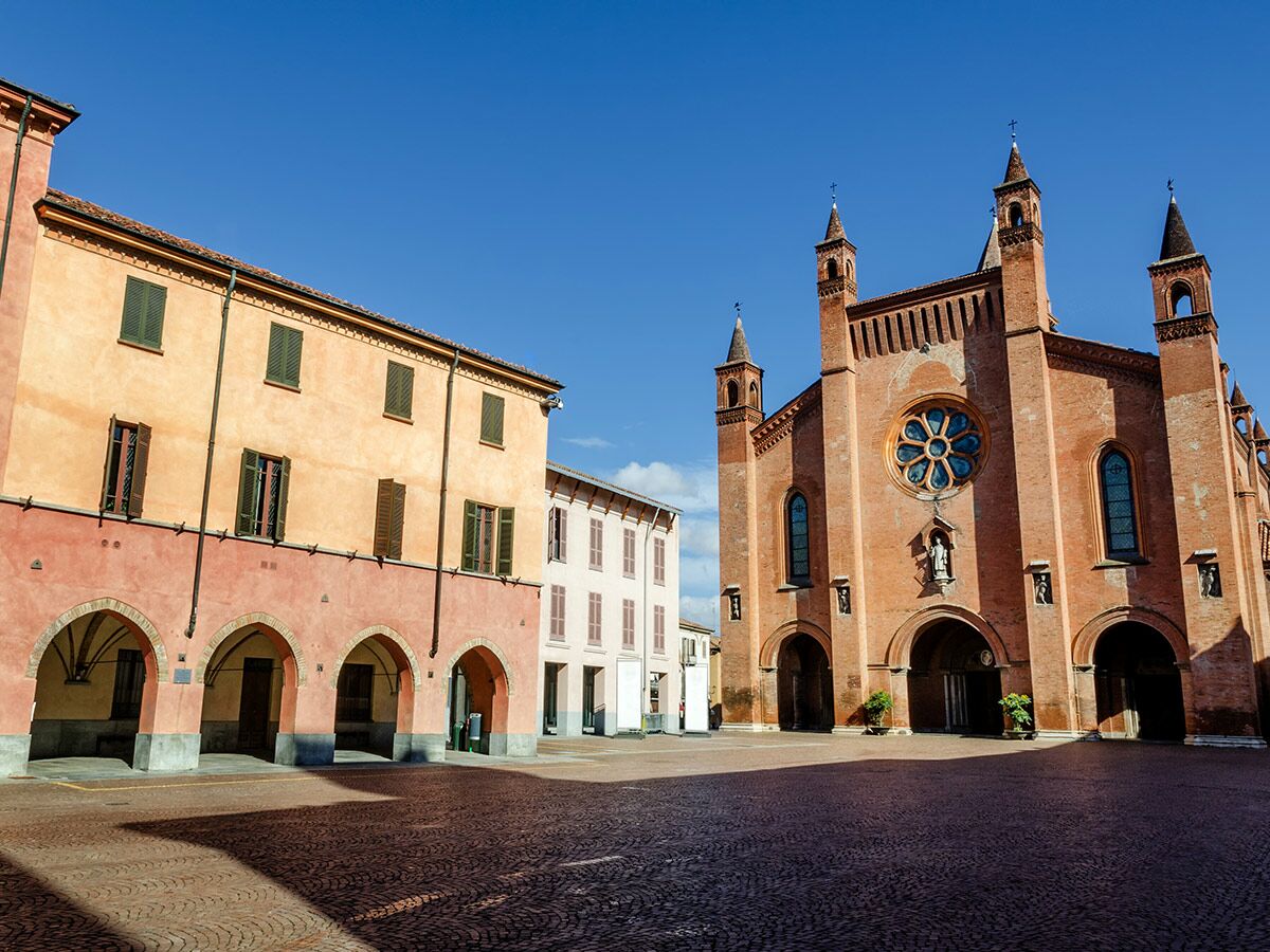 Historischer Platz in Alba, Italien, mit der Kathedrale San Lorenzo aus rotem Backstein und farbigen Gebäuden unter blauem Himmel.