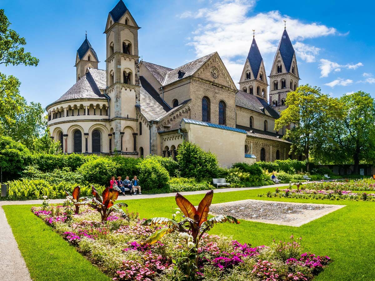 Blick auf eine historische romanische Basilika mit mehreren Türmen, umgeben von einem üppigen grünen Klostergarten mit Blumenbeeten unter blauem Himmel.