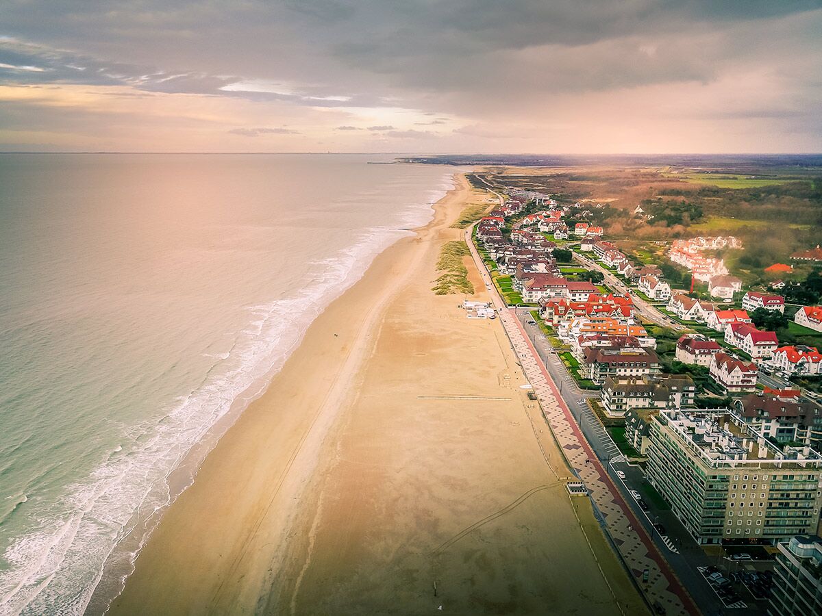Luftaufnahme einer weitläufigen Küstenlandschaft mit breitem Sandstrand, azurblauem Meer, Promenade und charmanten Häusern.