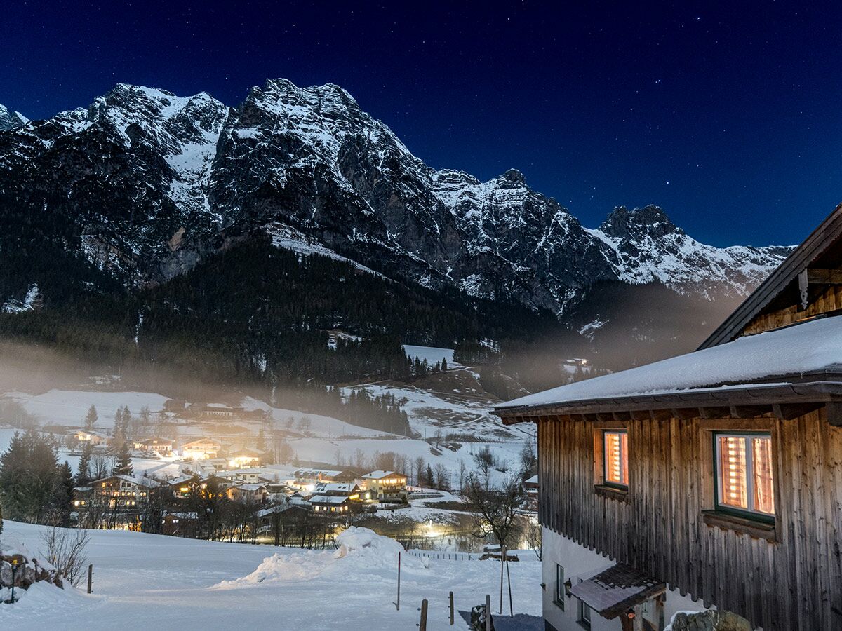 Verschneites Alpen-Bergdorf bei Nacht unter funkelndem Sternenhimmel, mit warm beleuchteten Häusern und imposanten Bergen.
