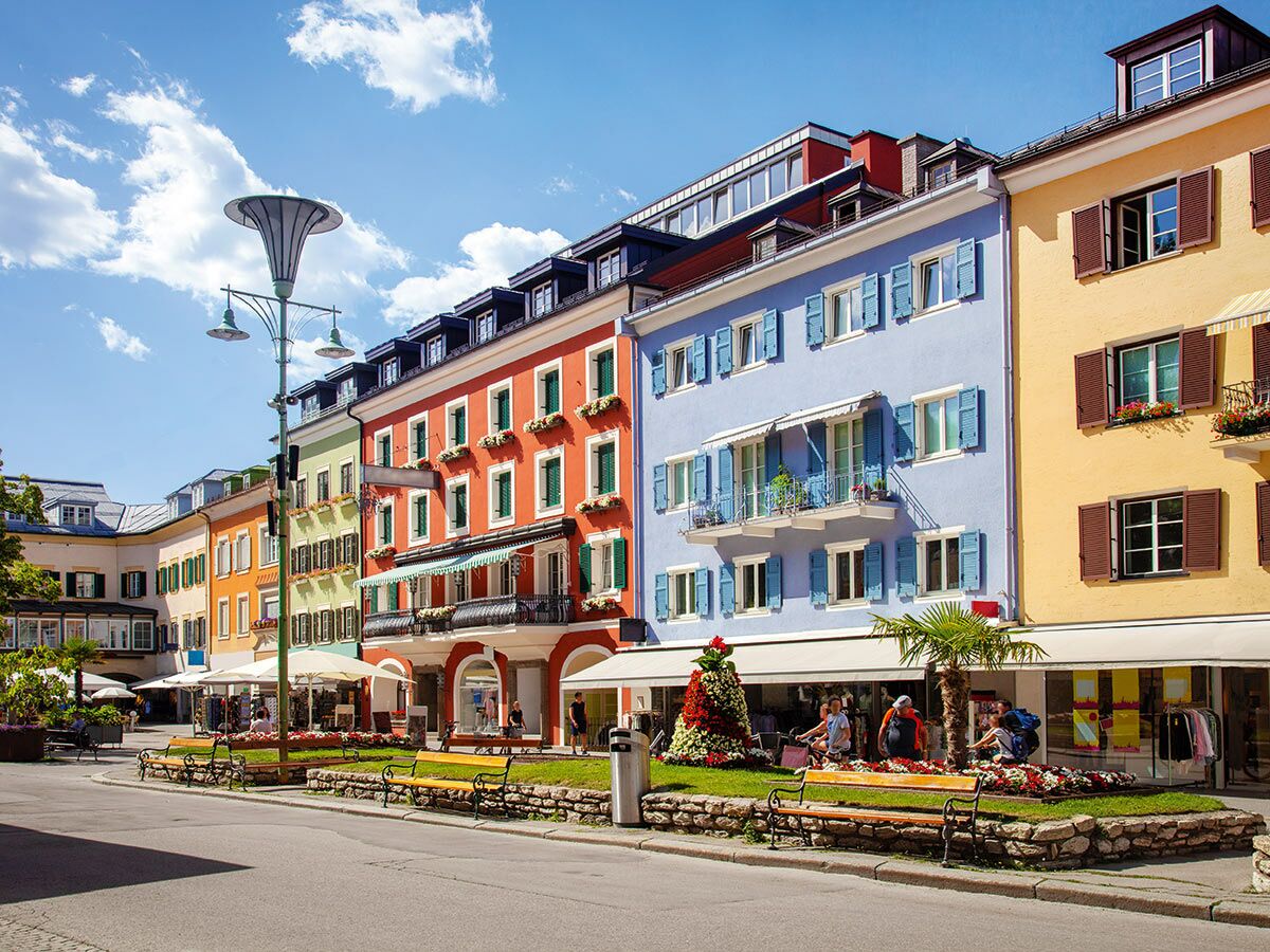 Blick auf eine bunte Straße mit traditionellen Gebäuden, Geschäften, Bänken und Blumen unter blauem Himmel.