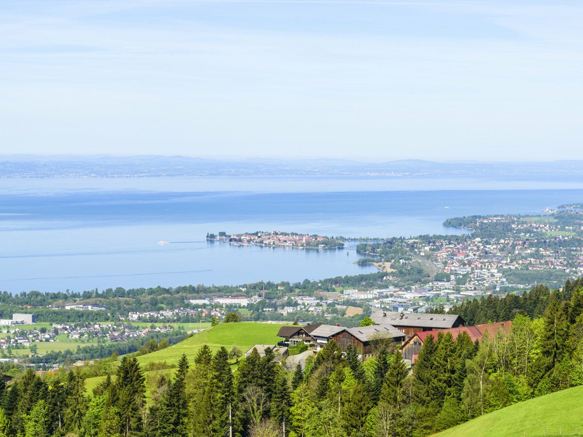 Panoramablick auf den Bodensee mit der Inselstadt Lindau, grünen Ufern, Wäldern und Häusern unter blauem Himmel.