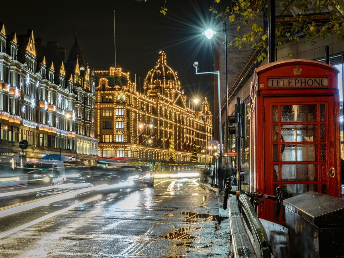 Beleuchtetes Harrods Kaufhaus und rote Telefonzelle in London bei Nacht auf nasser Straße mit Autolichtspuren.