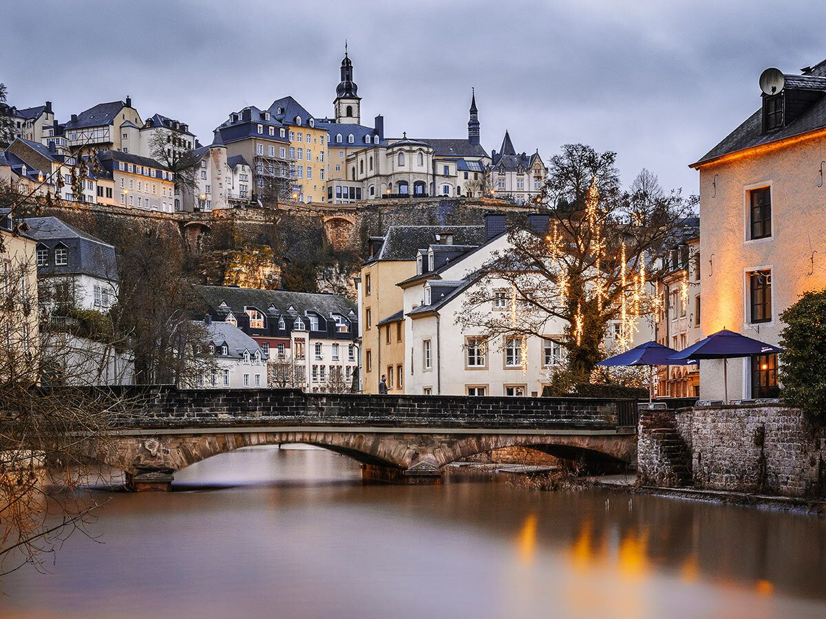 Historische Stadt Luxemburg: Grund-Viertel mit Fluss Alzette, alter Steinbrücke, beleuchteten Häusern, Kirche und Lichtern bei Abenddämmerung.