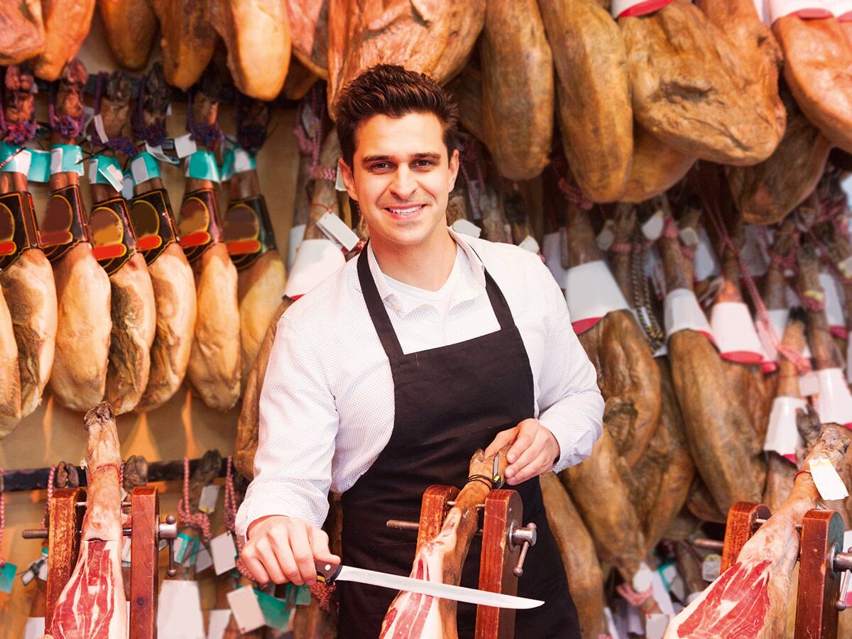 Lächelnder Metzger schneidet hauchdünne Scheiben von Jamon Serrano in einem traditionellen spanischen Schinkenladen mit vielen hängenden Schinken.