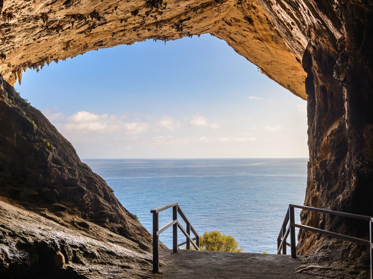 Blick aus einer großen Felsenhöhle auf das weite blaue Meer, den Himmel und eine Treppe mit Geländer am Ausgang.