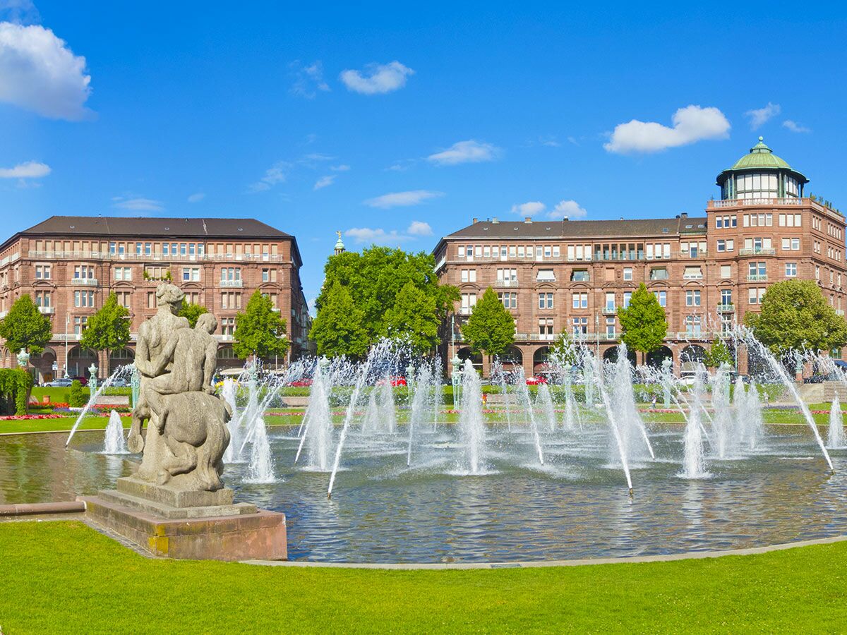 Großer Stadtbrunnen mit Wasserspielen, einer Skulptur und imposanten historischen Gebäuden unter blauem Himmel an einem sonnigen Tag.