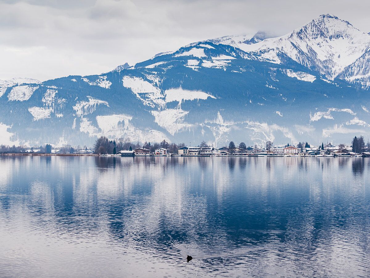 Malerische Winterlandschaft mit schneebedeckten Bergen, einem ruhigen See und einem Dorf am Ufer, das sich im Wasser spiegelt.
