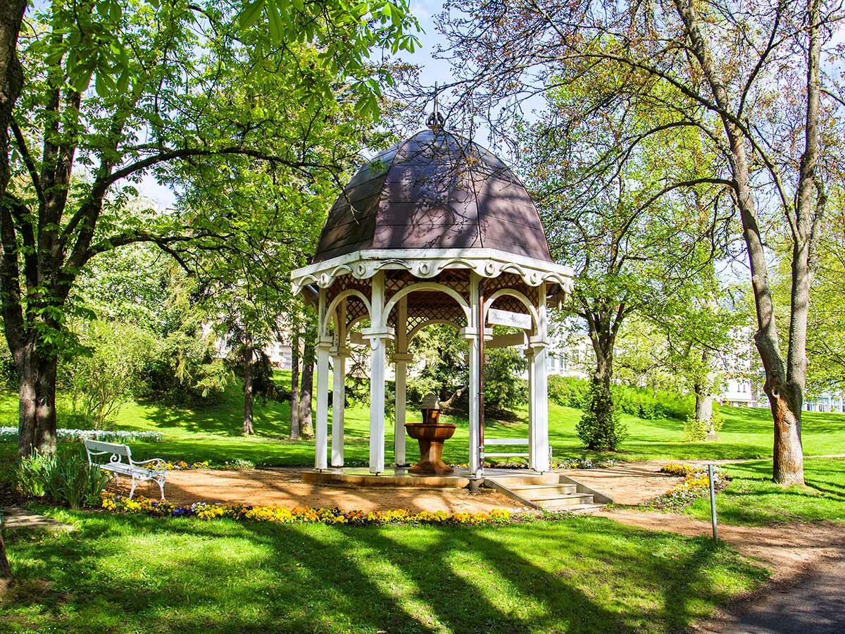 Eleganter Pavillon mit Brunnen in sonnigem Park, umgeben von Bäumen, Blumen und einer weißen Bank.