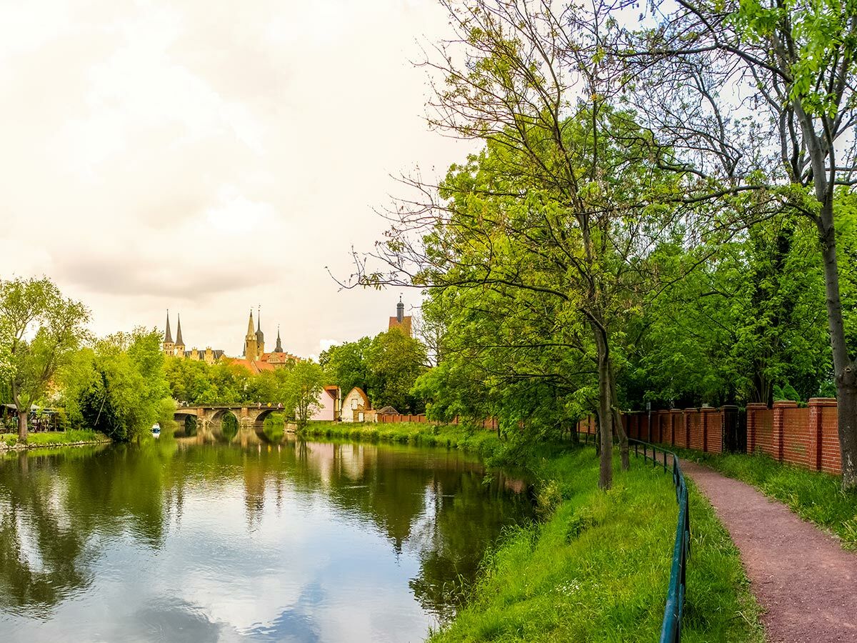 Malerische Altstadt mit Fluss, alter Brücke, grünen Bäumen und einem Spazierweg am Ufer.