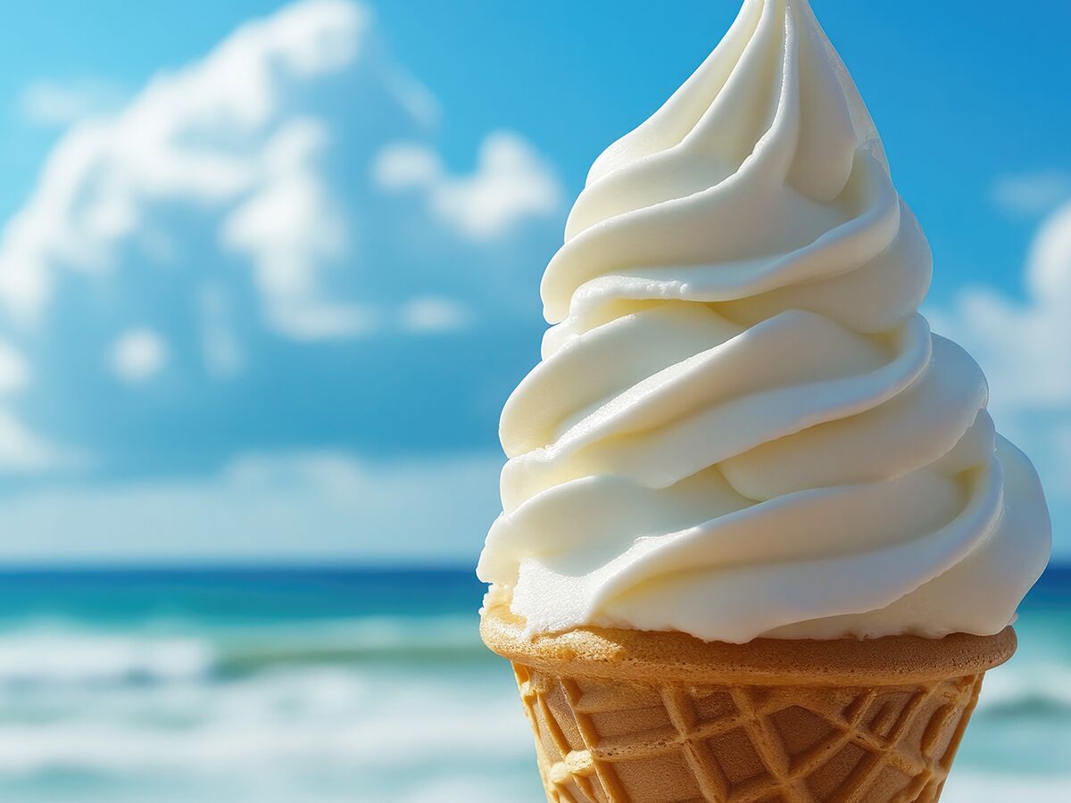 Nahaufnahme eines cremigen Vanille-Softeises in einer knusprigen Waffel vor unscharfem blauen Himmel und Meer.