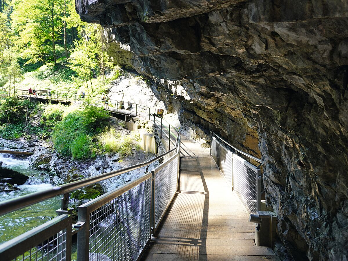 Felsiger Wanderweg mit Metallgeländer führt durch eine schattige Klamm mit reißendem Fluss und grünen Bäumen.