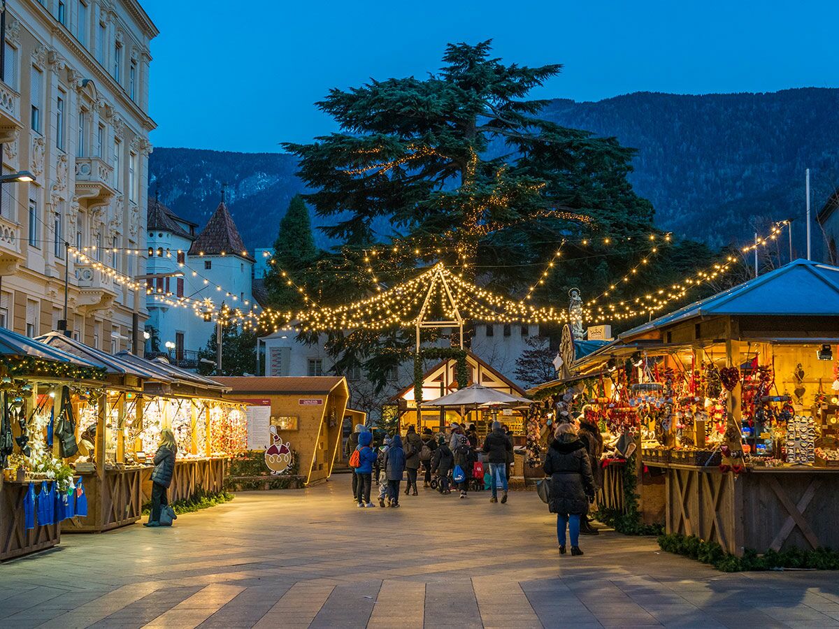 Weihnachtsmarkt in Meran bei Abenddämmerung mit festlicher Beleuchtung, geschmückten Buden und Besuchern, im Hintergrund Berge.