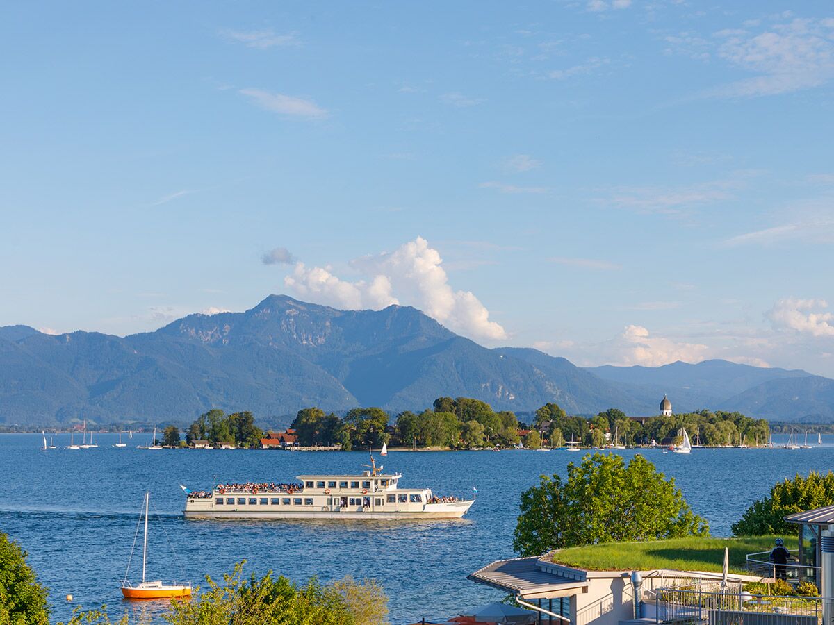Panoramablick auf bayerischen See mit Alpen im Hintergrund. Ein Passagierschiff, Segelboote, Insel und grüne Uferlandschaft sind zu sehen.