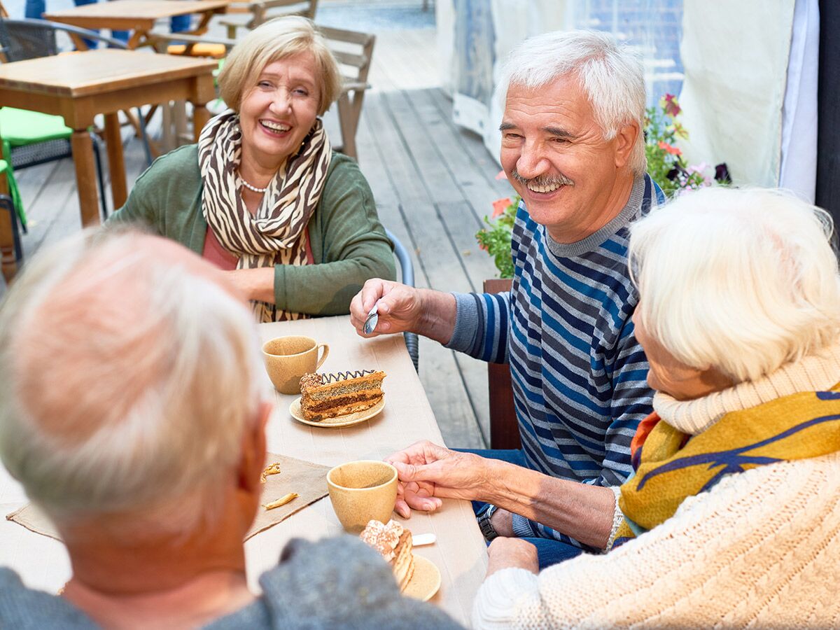 Glückliche Senioren sitzen an einem Cafétisch im Freien, trinken Kaffee und essen Kuchen, während sie sich unterhalten und lachen.