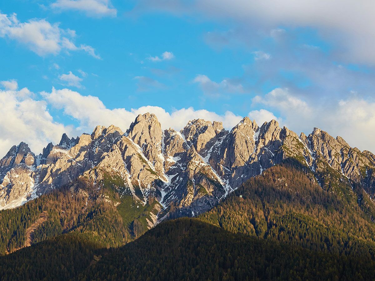 Majestätische felsige Berggipfel der Alpen, teilweise schneebedeckt, unter einem blauen Himmel mit Wolken und bewaldeten Hängen.