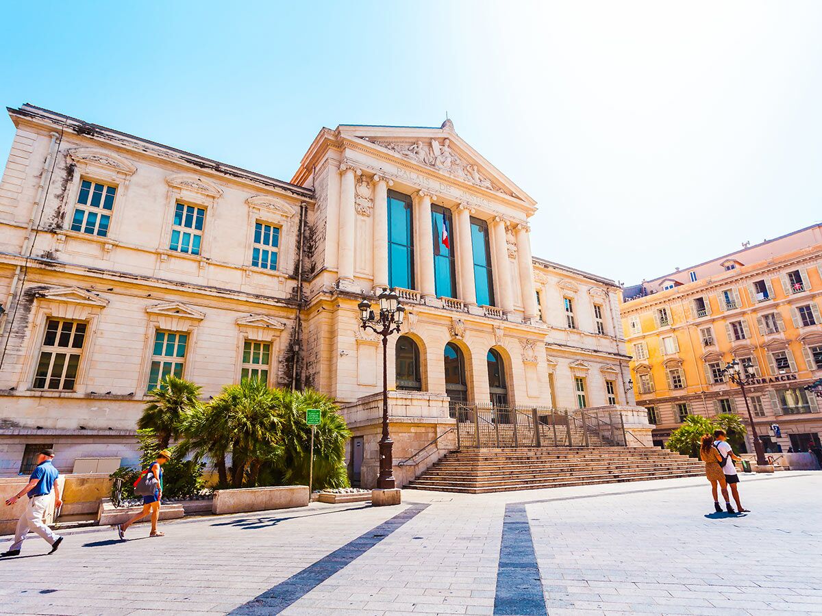 Der historische Palais de Justice in Nizza, Frankreich, mit Säulen, Treppen und Menschen bei sonnigem Wetter.