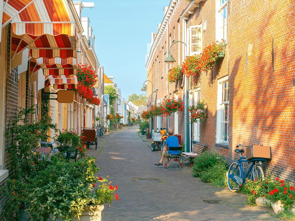 Schmale, sonnige Gasse mit historischen Backsteinhäusern, bunten Markisen und roten Blumen in Fensterkästen.