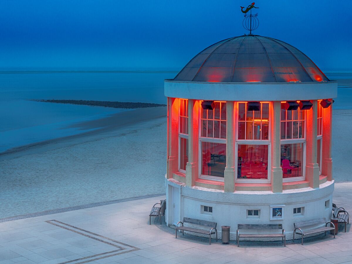 Roterleuchteter Strandpavillon mit Domdach und Meerjungfrauen-Wetterfahne bei blauem Abendhimmel am Strand.