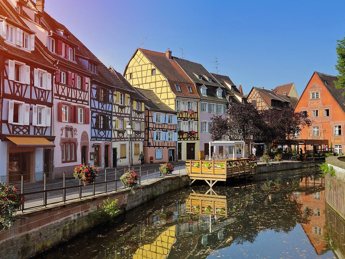 Bunte Fachwerkhäuser spiegeln sich im malerischen Kanal von Colmar, Elsass, unter blauem Himmel mit Sonnentouch.