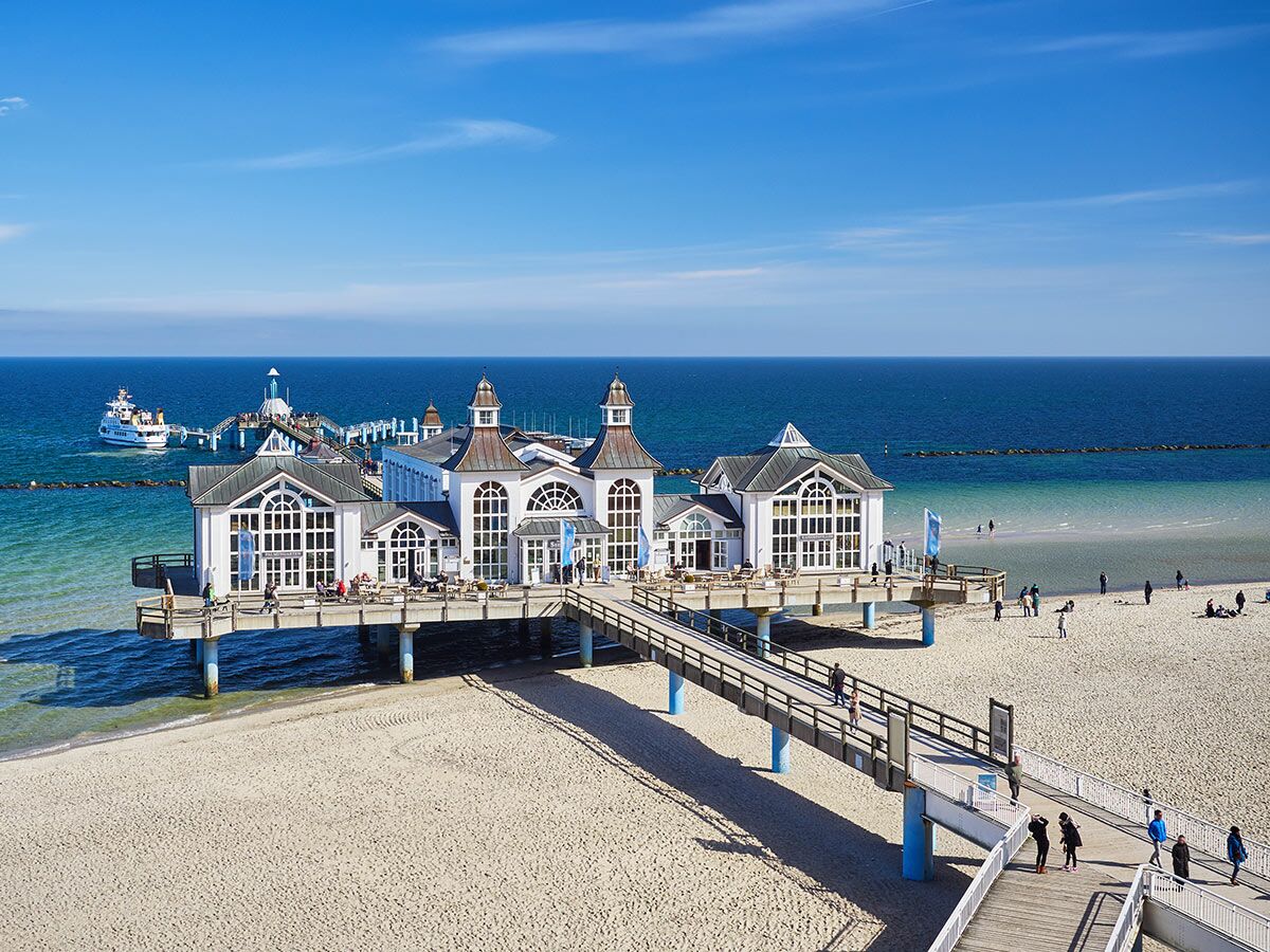 Panoramablick auf die Seebrücke Sellin auf Rügen mit weißen Gebäuden, sonnigem Strand, blauem Meer und Menschen.
