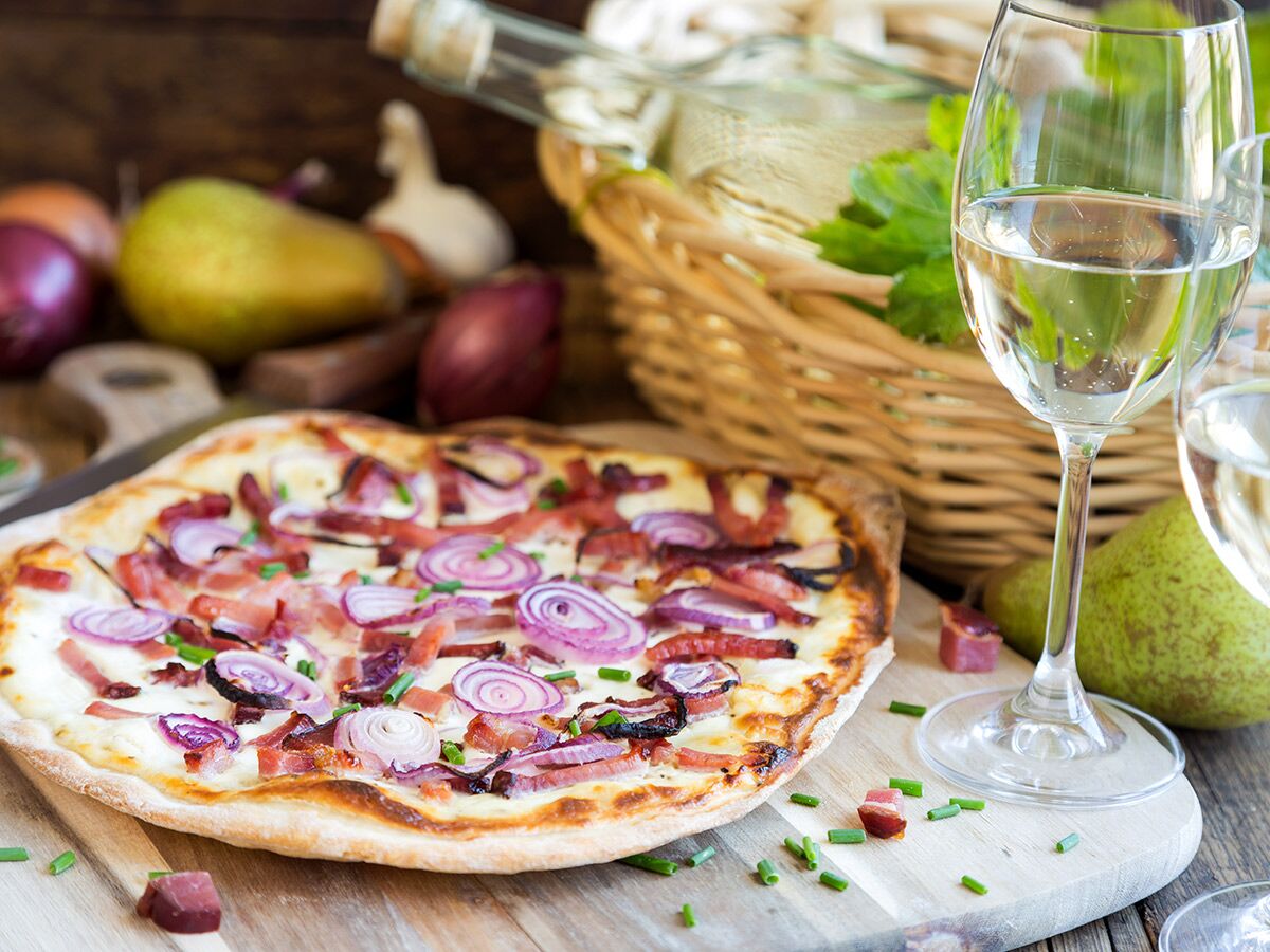Rustikaler Flammkuchen mit roten Zwiebeln und Speck auf Holzbrett, daneben Weißwein im Glas und Korb.