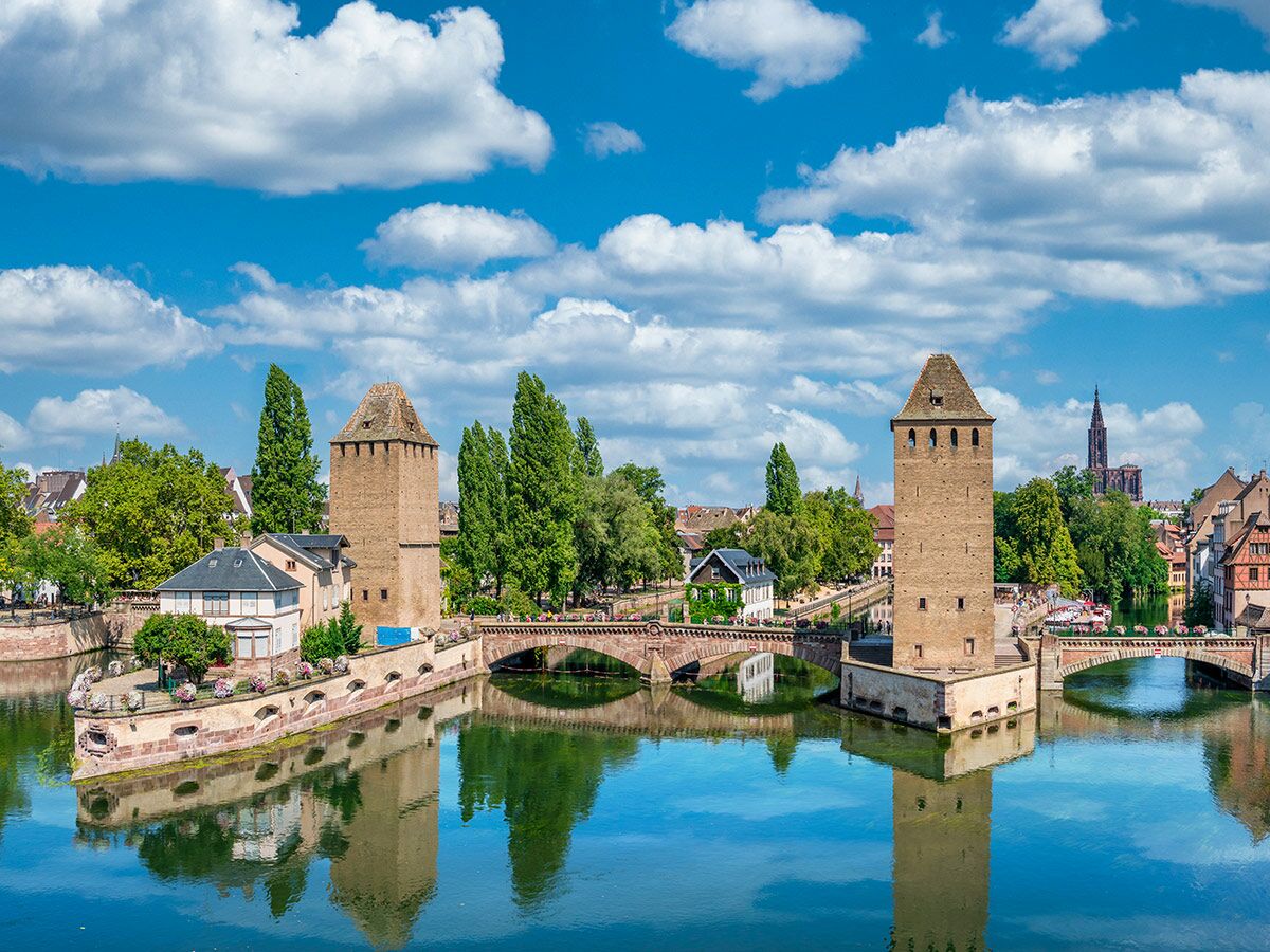 Historische Ponts Couverts Straßburg: Wehrtürme, Brücken und Fluss Ill spiegeln die grüne Umgebung und die Kathedrale unter blauem Himmel.