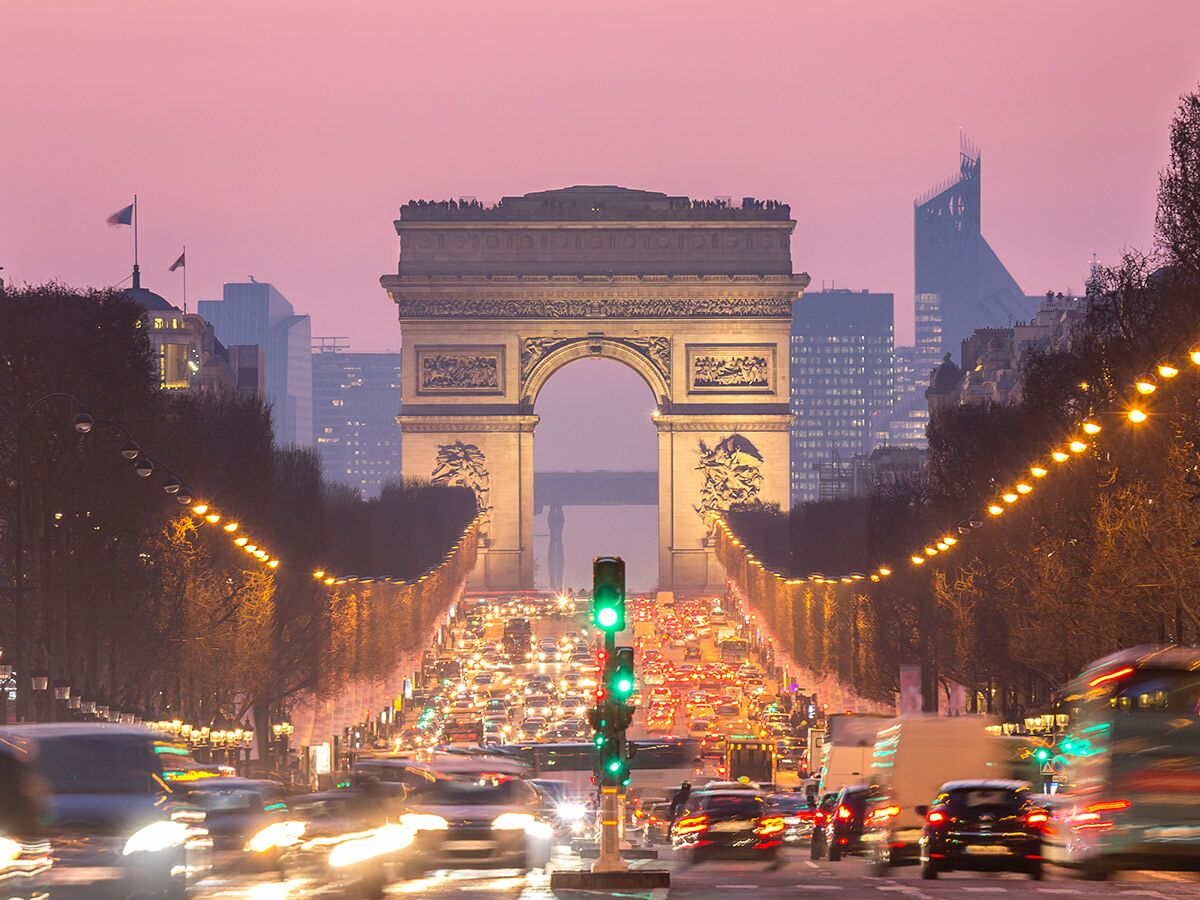 Arc de Triomphe in Paris bei Sonnenuntergang, beleuchtete Champs-Élysées mit starkem Verkehr und rosa Himmel.