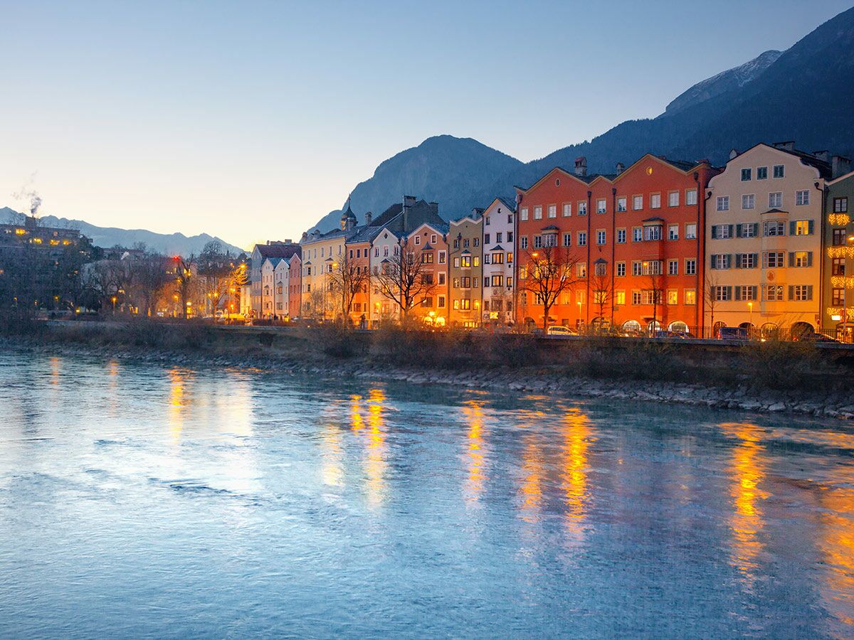 Bunte Häuser der Innsbrucker Altstadt am Fluss Inn in der Abenddämmerung, mit Lichtern, die sich im Wasser spiegeln und Alpenbergen im Hintergrund.