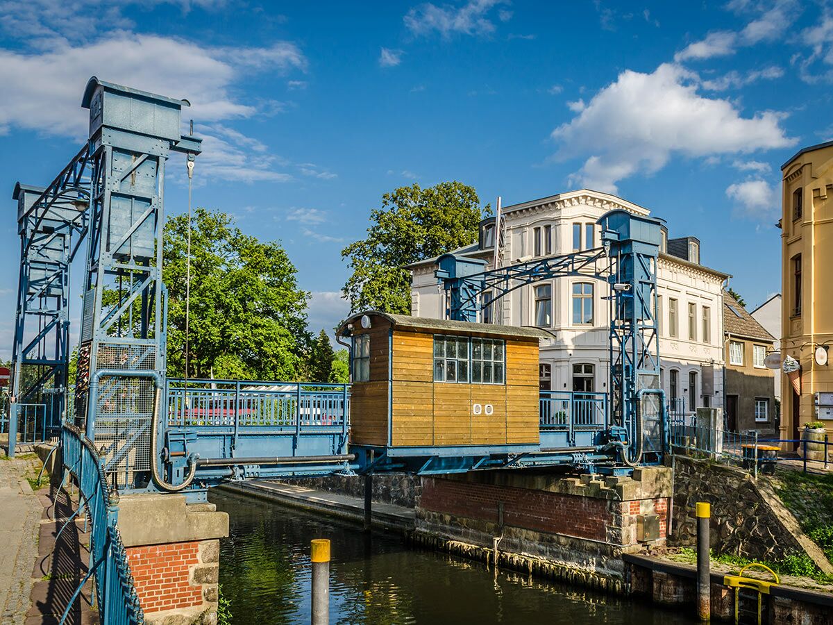 Historische blaue Klappbrücke mit Holzführerhaus über einen Kanal, umgeben von Bäumen und alten Gebäuden unter blauem Himmel.