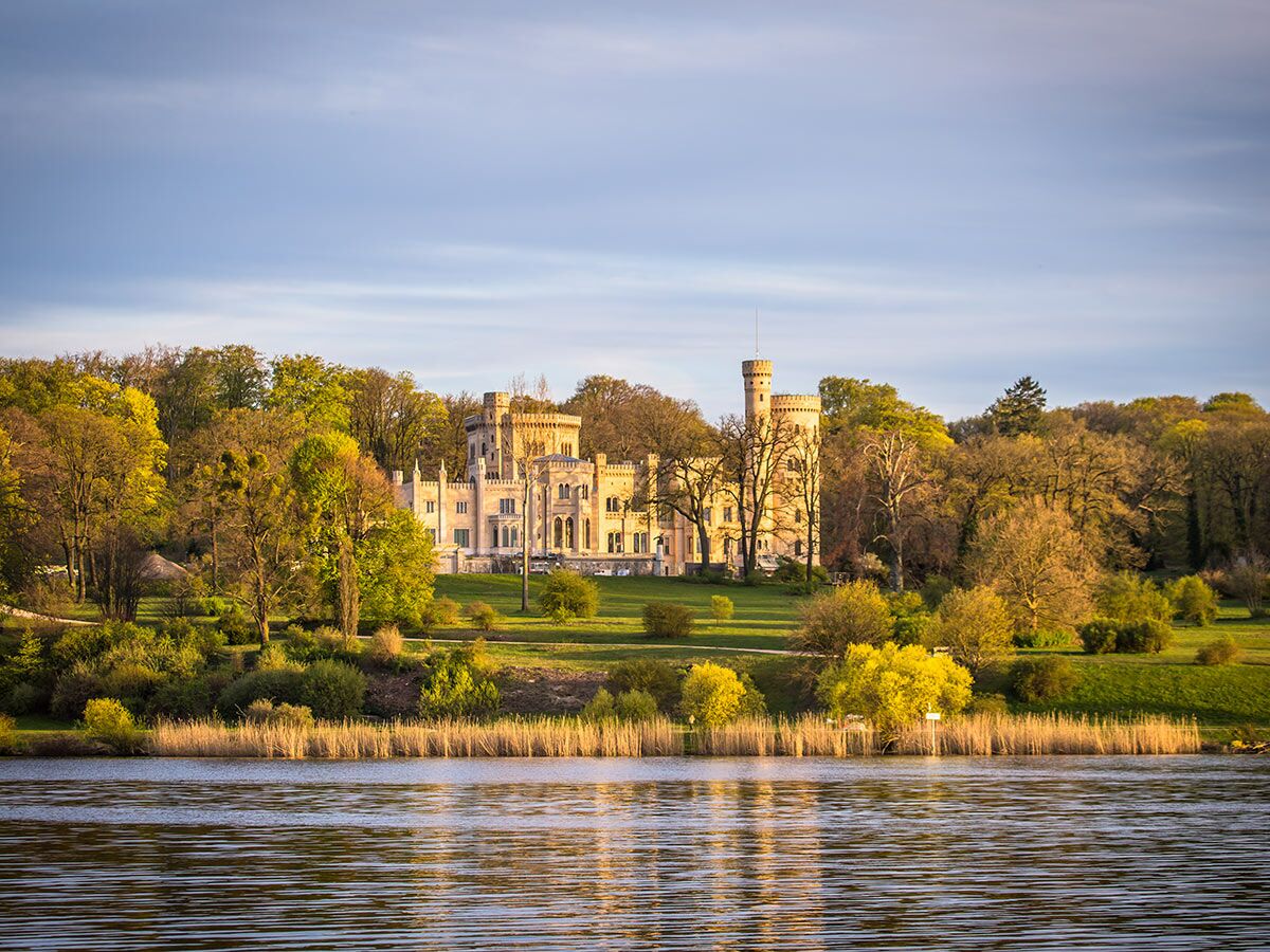 Historisches Schloss Babelsberg in Potsdam, von Seeufer aus gesehen, umgeben von grünen Bäumen im goldenen Sonnenlicht.