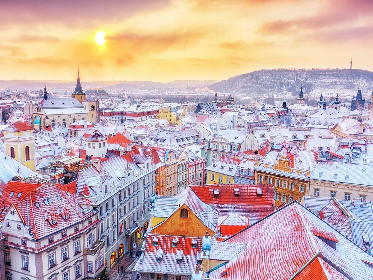 Panorama von Prag im Winter bei Sonnenuntergang, mit Schnee bedeckten roten Dächern der Altstadt, historischen Gebäuden und einem Hügel im Hintergrund.