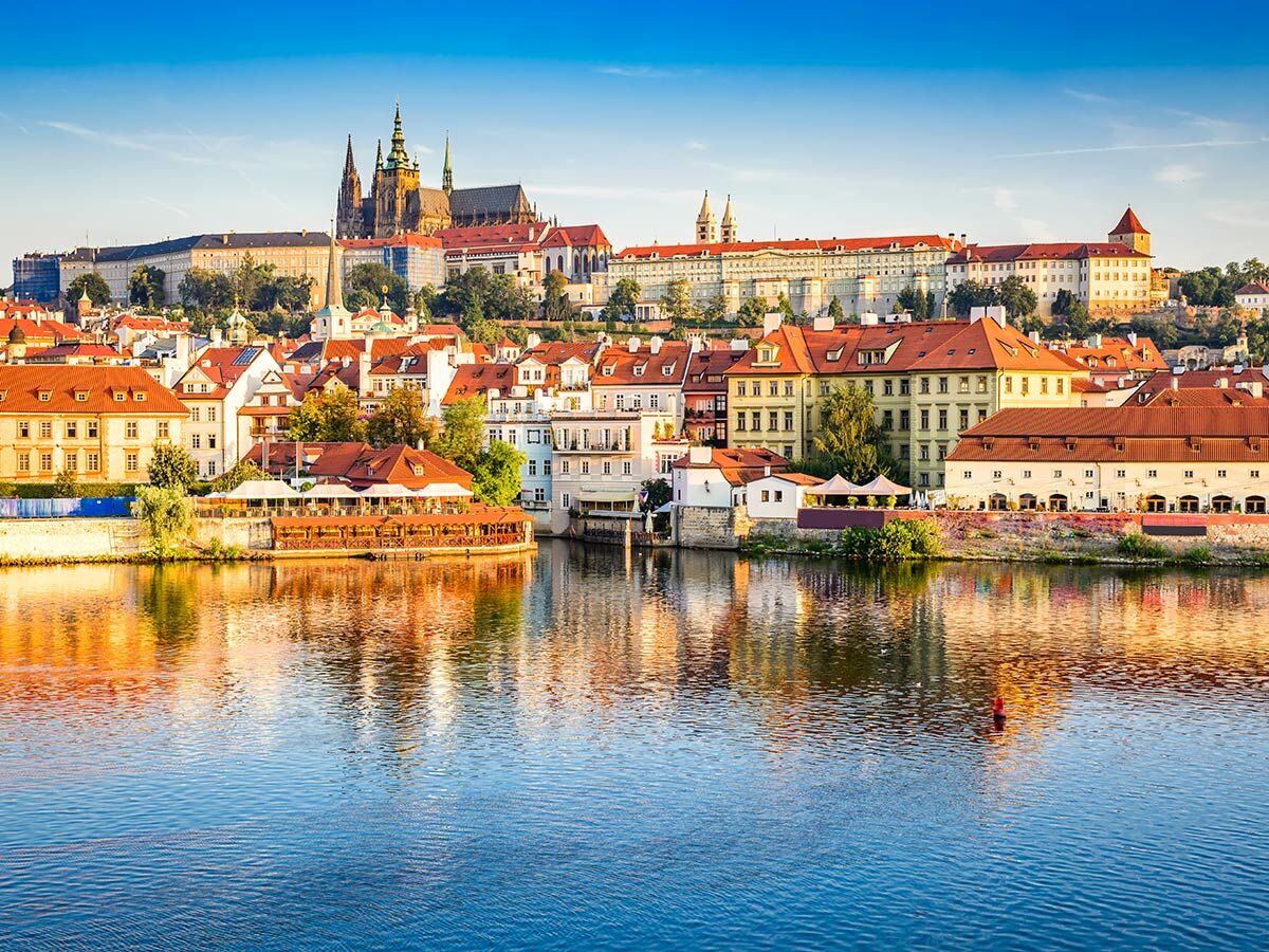 Historische Stadtansicht von Prag mit der Moldau, roten Dächern und dem Prager Schloss unter blauem Himmel.