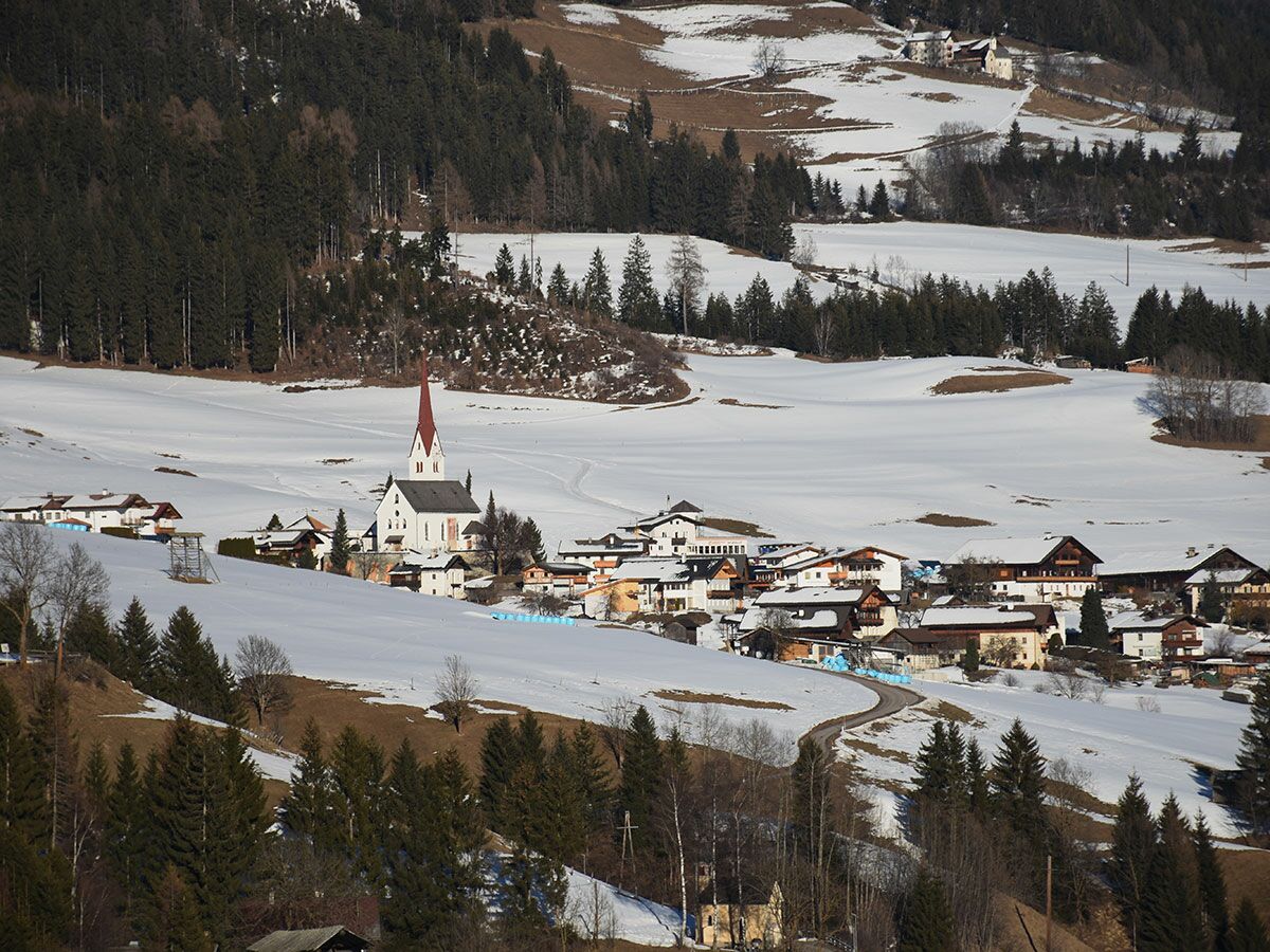 Malerisches Winterdorf in den Alpen mit weißer Kirche, roten Dachspitzen, traditionellen Häusern und verschneiten Feldern.