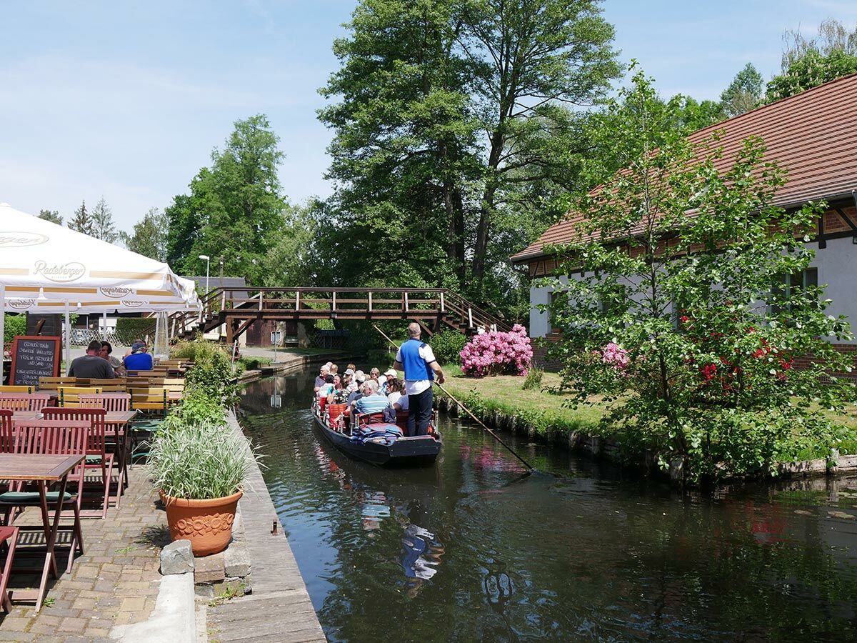 Besucher genießen Kahnfahrt im Spreewald, vorbeifahrend an einem Restaurant mit Biergarten und Holzbrücke.