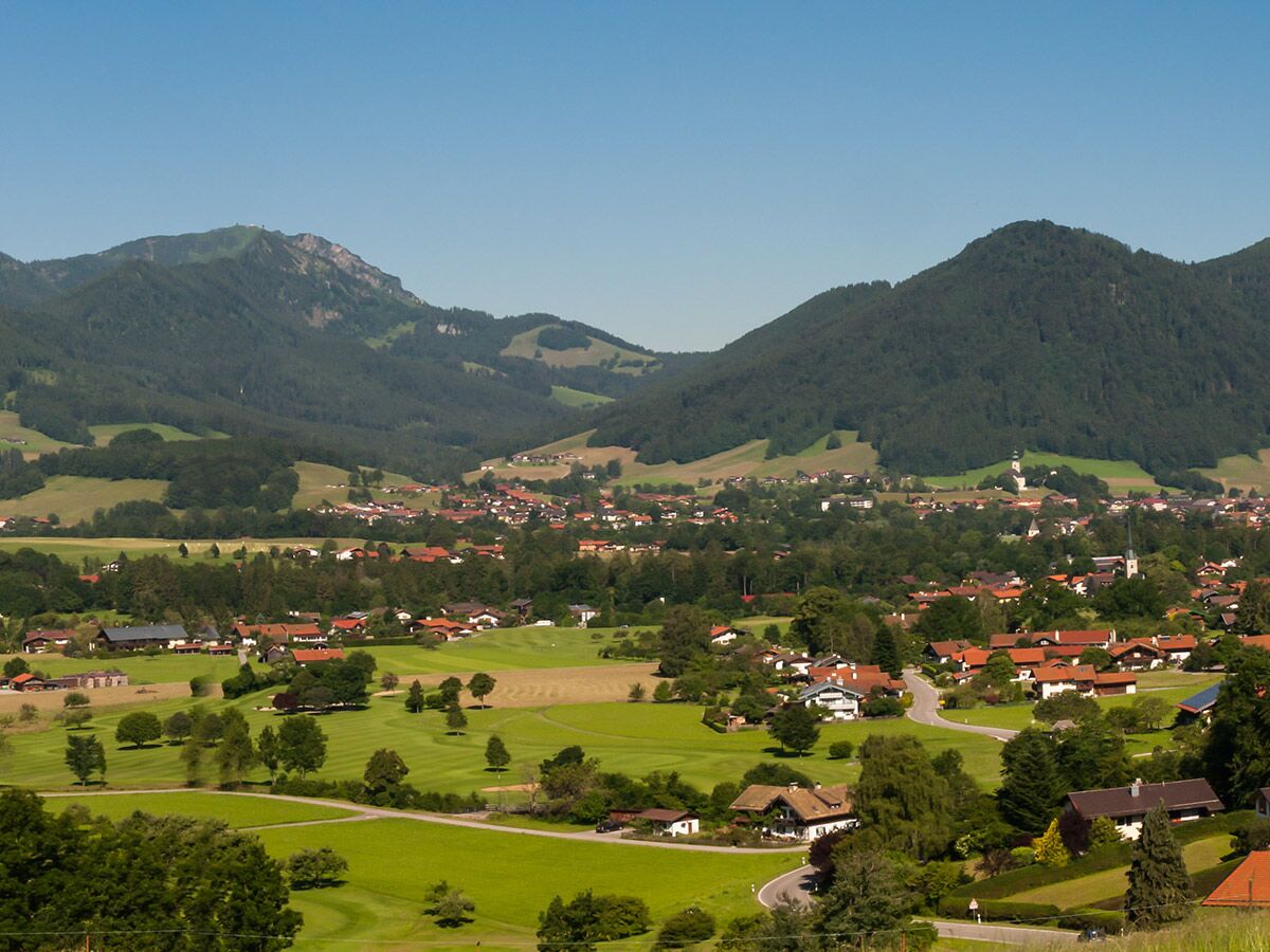 Idyllisches Dorf in einem grünen Tal, umgeben von bewaldeten Bergen und einem Golfplatz unter blauem Himmel.