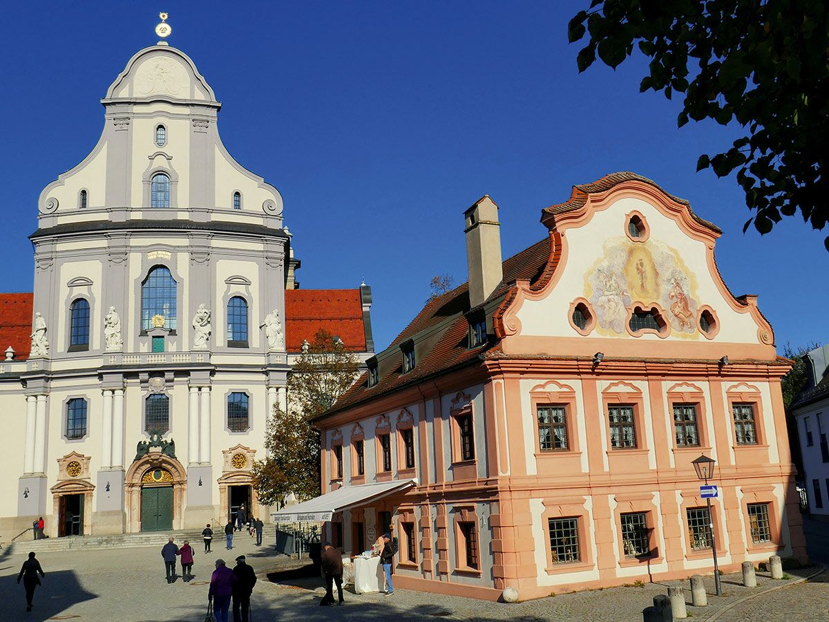 Die barocke St. Michael Kirche und ein Rokoko-Gebäude mit Fresko auf einem sonnigen Platz in München. Menschen gehen umher.