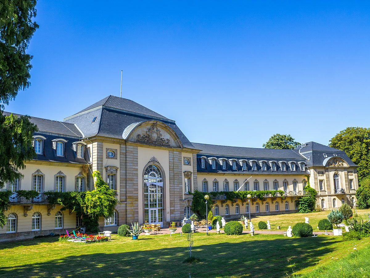 Elegantes historisches Schloss oder Hotelgebäude mit gepflegtem Garten, Rasen und Statuen unter blauem Himmel an einem sonnigen Tag.