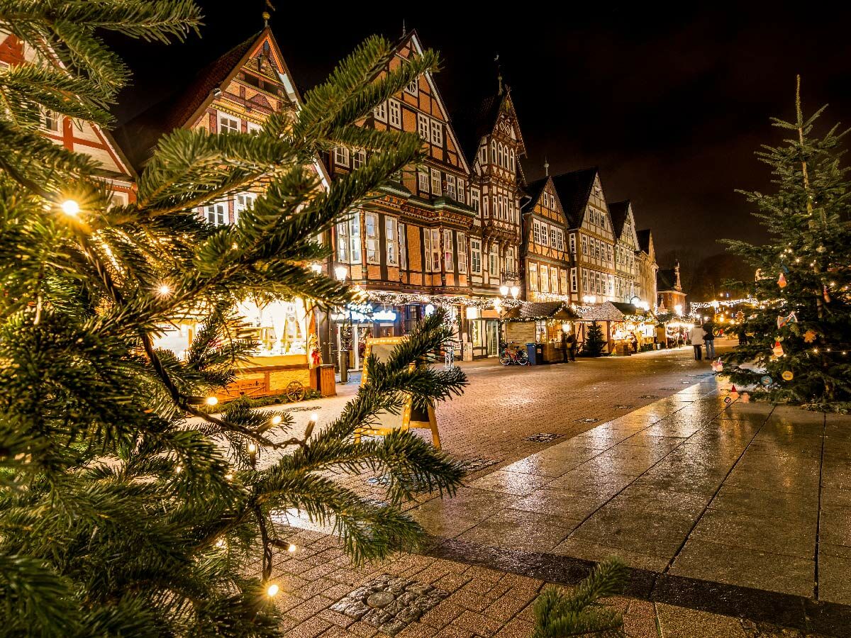 Festlicher Weihnachtsmarkt mit geschmückten Fachwerkhäusern, beleuchteten Ständen und Tannenbäumen bei Nacht, spiegelnd auf nassem Kopfsteinpflaster.