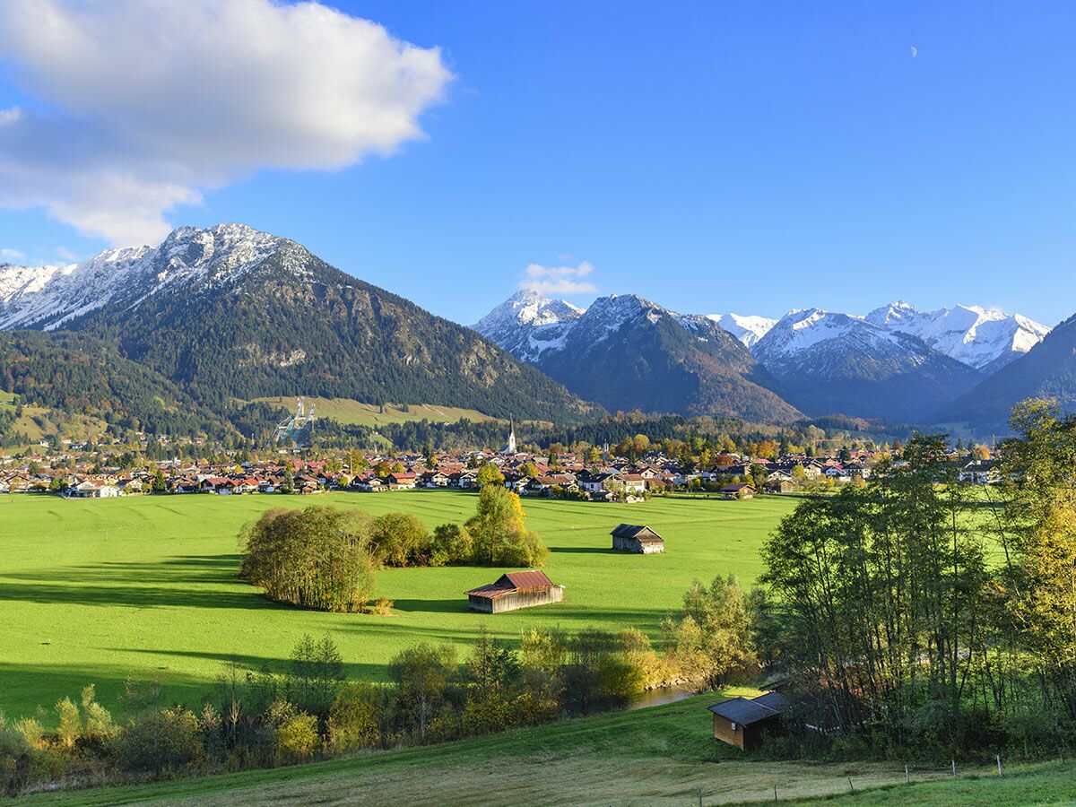 Malerische Alpenlandschaft im Herbst mit schneebedeckten Bergen, grünem Tal, einem bayerischen Dorf und einer Skisprungschanze.